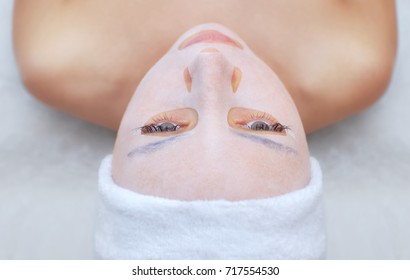 The cosmetologist for procedure of cleansing and moisturizing the skin, applying a sheet mask to the face of a young woman in beauty salon.Cosmetology and professional skin care.