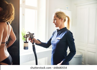 Cosmetologist using aerograph for spray tan apply to young female back wearing bikini in beauty salon. - Shutterstock ID 648680554