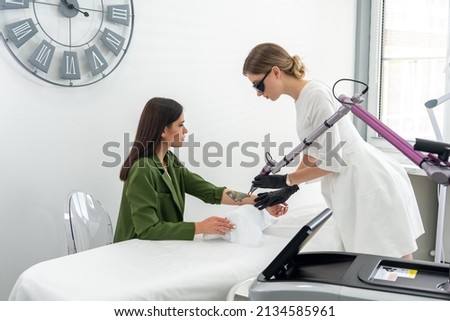 Cosmetologist using laser device to remove an unwanted tattoo from female arm. Concept of erasing tattoos as an expensive procedure in a cosmetology clinic