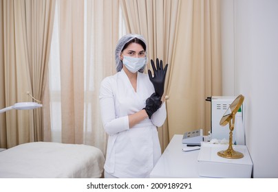 Cosmetologist In A White Uniform Puts On Black Disposable Gloves In The Office