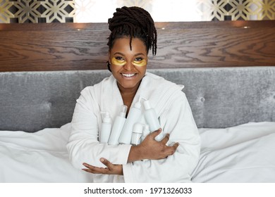 cosmetology and dermatology. happy african woman sitting on bed at home, using cream, holding cosmetics bottles tubes