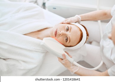 Cosmetology RF lifting equipment. Anti-age and anti-wrinkle apatherapy. Top view of beautiful woman in white bathrobe getting facial treatment at Cosmetology Salon.