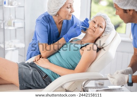 In cosmetology salon, elderly woman doctor and male assistant perform rejuvenation procedure for female patient, gives injection of botulinum toxin in lip area
