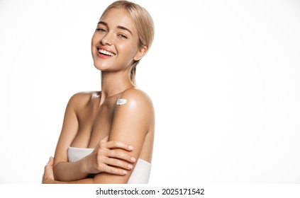 Cosmetology and spa. Healthy young blond woman wear towel after shower, smiling and looking happy, using body lotion, moisturizer cream on shoulder, apply skincare products, white background