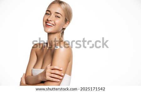 Cosmetology and spa. Healthy young blond woman wear towel after shower, smiling and looking happy, using body lotion, moisturizer cream on shoulder, apply skincare products, white background