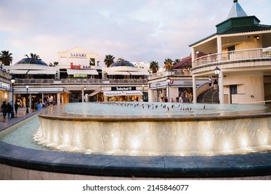 Costa Adeje, Spain - March 10, 2022: 
 Outdoor shopping mall with many famous brands store on background. Fountain close-up during evening dusk light.
