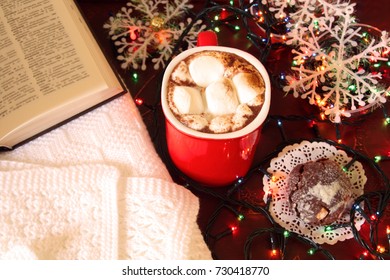 Cosy Christmas Eve Scene: Winter Hot Spicy Drink Cacao With Marshmallows, Open Book And Christmas Chocolate Cookies On Background Of White Blanket, Electric Lights And White Snowflakes