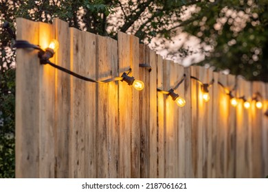 Cosy light bulbs lined up in a row, against a wooden garden fence. There are some green bushes and green grass in the background. It's a sunny Summer day evening at a country wedding or other - Powered by Shutterstock