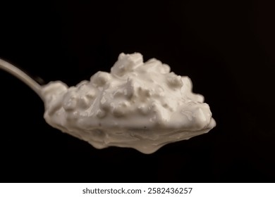 cottage cheese made from milk closeup, grainy cottage cheese with cream on the table, in a metal spoon on a black background