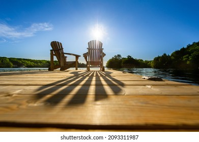 Cottage life - Sunrise on two empty Adirondack chairs sitting on a dock on a lake in Muskoka, Ontario Canada. The sun rays create long shadows on the wooden pier.