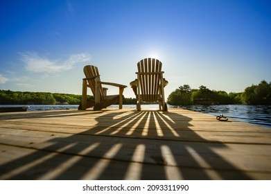 Cottage life - Sunrise on two empty Adirondack chairs sitting on a dock on a lake in Muskoka, Ontario Canada. The sun rays create long shadows on the wooden pier. - Shutterstock ID 2093311990