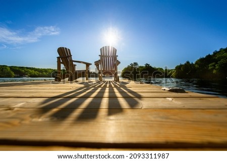 Cottage life - Sunrise on two empty Adirondack chairs sitting on a dock on a lake in Muskoka, Ontario Canada. The sun rays create long shadows on the wooden pier.