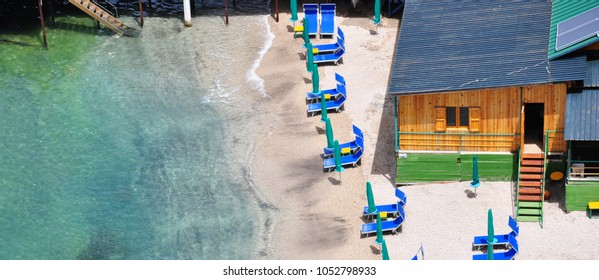 Cottage On A Sand Beach With Blue Sun Beds And Umbrellas On Capri Island. Half Left Of The Photo Is The Blue Transparent Sea Water On Shore With Line Waves, Top View