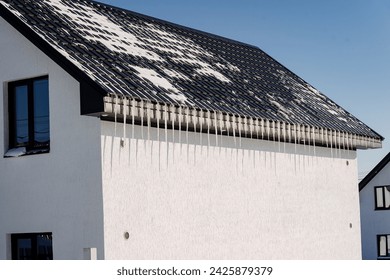 A cottagestyle house with a white exterior and a contrasting black roof covered in snow, blending in with the winter sky - Powered by Shutterstock