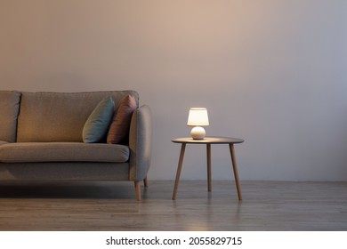 Couch With Pillows, Glowing Lamp On Table At Home On Gray Wall Background In Evening In Living Room. Advertising Of Modern Minimalist Interior, Comfortable Furniture And Real Estate Blog, Free Space