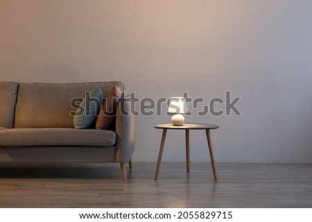 Couch with pillows, glowing lamp on table at home on gray wall background in evening in living room. Advertising of modern minimalist interior, comfortable furniture and real estate blog, free space