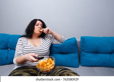 Couch potato overweight woman sitting on the sofa, eating chips while watching TV. Sedentary lifestyle, bad habits, eating disorder concept 