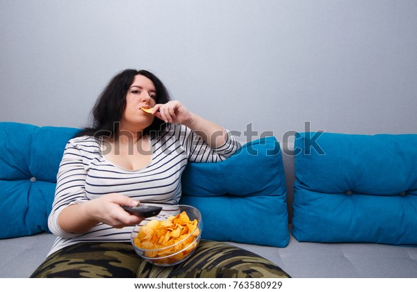 Couch potato overweight woman sitting on the\
sofa, eating chips while watching TV. Sedentary lifestyle, bad\
habits, eating disorder concept\

