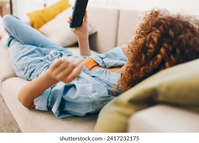 Couch Surfer: A Happy Young Woman Holding a Smartphone, Engaging in Online Games and Chatting, Enjoying Leisure Time at Home, in her Cozy Living Room - Powered by Shutterstock