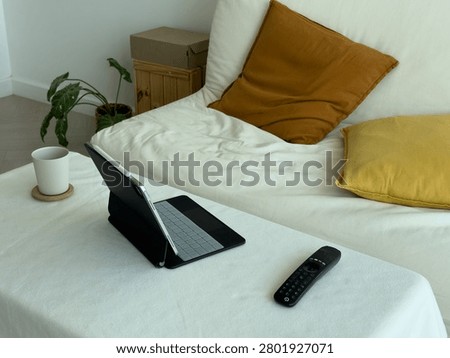 Couch workspace with tablet and remote on soft mattress and mustard pillows, open folio and coffee mug beside a leafy plant