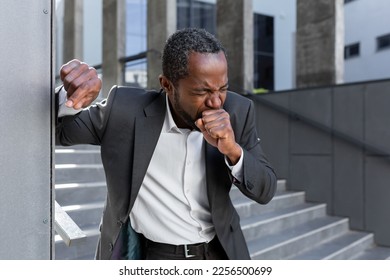 Cough attack. An African-American businessman in a suit is holding on to the office wall outside, coughing hard, covering his mouth with his hand. feels chest pain. - Powered by Shutterstock