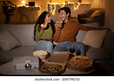 Coule Eating Pizza Talking And Watching Movie On Projector Sitting On Sofa In Living Room Indoor At Night, Having Domestic Date. Spouses Having Fun Watching Films And Enjoying Junk Food Together