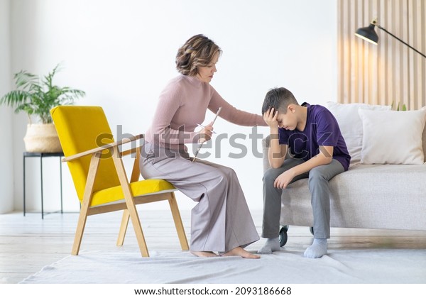 Counseling\
adolescent psychologist calms and holds adolescent by shoulder. Boy\
in purple t-shirt is worried and holds his head with his hand,\
while sitting on couch in minimalistic\
room.