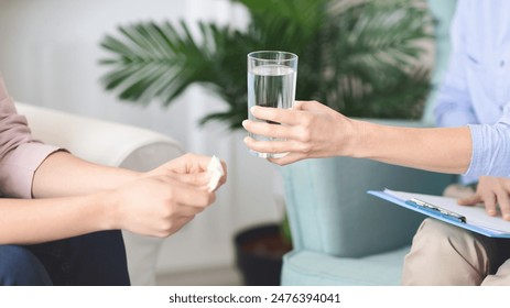 A counselor extends a glass of water to a seated individual holding a tissue, suggesting a supportive and comforting atmosphere in an office setting, cropped - Powered by Shutterstock