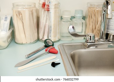 Counter With Sink In Doctors Office With Various Tools, And Mild Blurred Medical Supplies In Background. Includes Tongue Depressors, Plexors Or Percussor Swabs, 