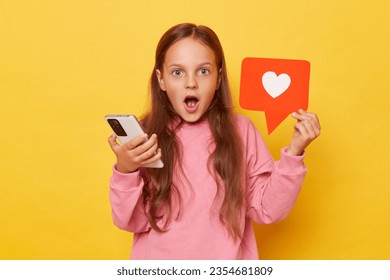 Counting clicks on viral content. Following a popular vlog on the web. Amazed shocked little girl wearing pink sweatshirt holding blogger heart like icon and phone isolated over yellow background