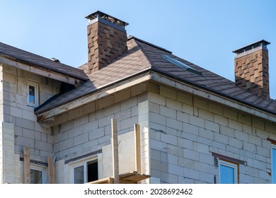 Country Cottage Made Of Porous Cement Block Under Construction With Finished Soft Roof With Chimney And Attic Window. Building With Scaffolding For Finishing Facade With Ceramic Tiles