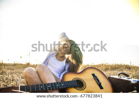 Country hippie girl with guitar at wheat field near old retro suitcase. Cute woman sit against sunny sky with sun and clouds. sun rays light. vintage case. sunny day light. 