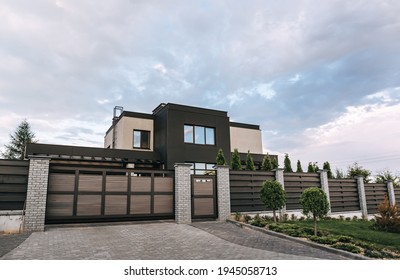 Country house with a flat roof and large windows in brown and beige colors. Exterior. Entrance group: gate and wicket. Fence: brick posts with sections of horizontal wooden planks. Landscaping design.