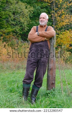 In a country house style with dungarees, sweater and rubber boots, an older man leans against a wooden post in the meadow in front of the colorful autumn forest and enjoys the beautiful view.
