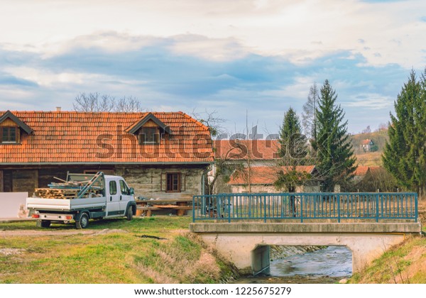 Country landscape: wooden house with a tiled\
roof, a car and a bridge over the\
stream
