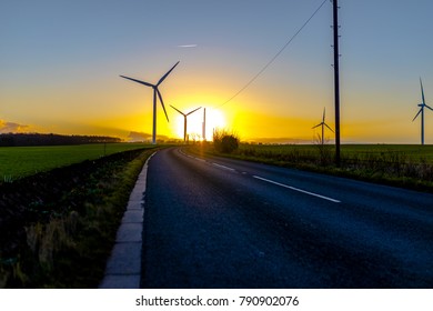 Country Road Leading To Turbines In A Field In The UK At Sunset Or Sunrise Against A Clear Winter Sky