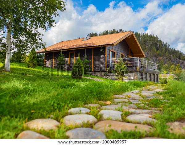 Country\
vacation in summer. Cottage near pine forest and rock. Cottages for\
rent concept. Suburban cottage vacation in nature. Stone path next\
to cottage with terrace. Suburban rental\
houses.