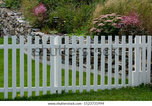 A country wooden white picket\
fence and gate divides gardens. There\'s a rock wall with pink\
flowers and shrubs on one side and green grass on the other side.\
