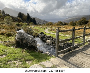 A countryside scene with an old wooden bridge over a small river and mountains in the distance. 