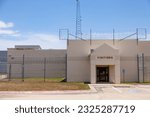 A county or city jail exterior on a sunny day with a blue sky and clouds shows a prison yard protected by barbed wire and a chain link fence.  City correctional facility near the visitor