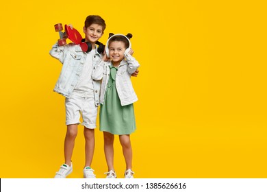 Couple of adorable stylish children with skateboard smiling and listening to music in headphones while standing against bright yellow background. Brother and sister hugging
