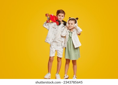 Couple of adorable stylish children with skateboard smiling and listening to music in headphones while standing against bright yellow background. Brother and sister hugging - Powered by Shutterstock