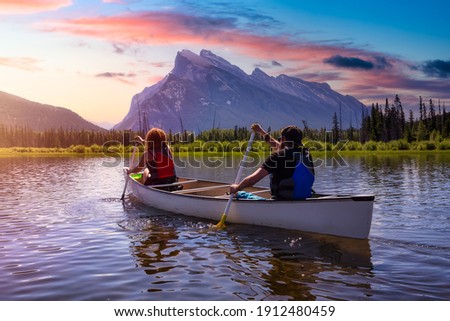 Couple adventurous friends are canoeing in a lake surrounded by the Canadian Mountains. Colorful Sunrise Sky Art Render. Taken in Vermilion Lakes, Banff, Alberta, Canada.