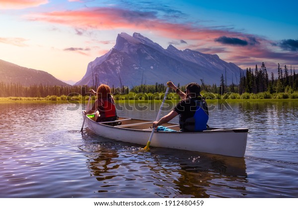 Couple\
adventurous friends are canoeing in a lake surrounded by the\
Canadian Mountains. Colorful Sunrise Sky Art Render. Taken in\
Vermilion Lakes, Banff, Alberta,\
Canada.