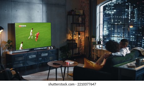 Couple Of African American Soccer Fans Relax On A Couch, Watch A Sports Match At Night At Home In Stylish Loft Apartment. Young Man And Woman Enjoying The Game Of Favorite Football Club.