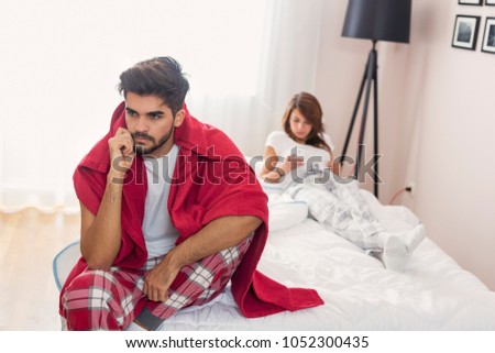 Couple after a fight in bedroom; man sitting on the edge of a bed angry while woman lying in the background with a smart phone