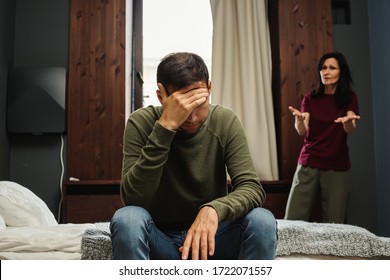 Couple with age gap or mother and adult son arguing in bedroom. Anxious middle aged woman blaming depressed young man sitting on bed and covering face with hand
