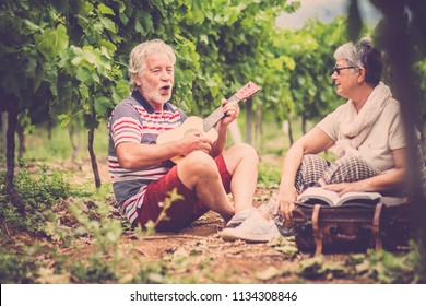 Couple Of Alternative Aged Older Traveler Stay Sitting Down In A Vineyard With The Luggage And Playing An Ukulele Acoustic Guitar And Reading A Paper Book. Easy Lifestyle In Outdoor Places And Nature