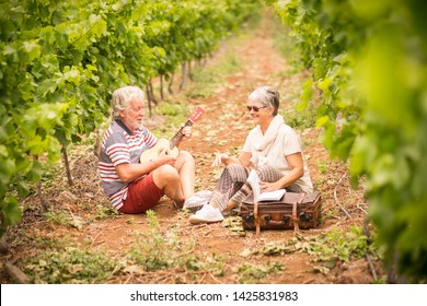 Couple Of Alternative Aged Older Traveler Stay Sitting Down In A Vineyard With The Luggage And Playing An Ukulele Acoustic Guitar And Reading A Paper Book. Easy Lifestyle In Outdoor Places And Nature