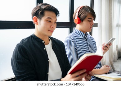 Couple of asian LGBTQ+ men spending time reading a book and listing music with wireless headphone and smartphone.